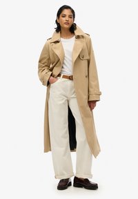 Beżowy płaszcz trencherowy z dużym kołnierzem i podwójnymi guzikami, noszony na białej koszuli. Luźno dopasowane białe spodnie i ciemne loafersy.