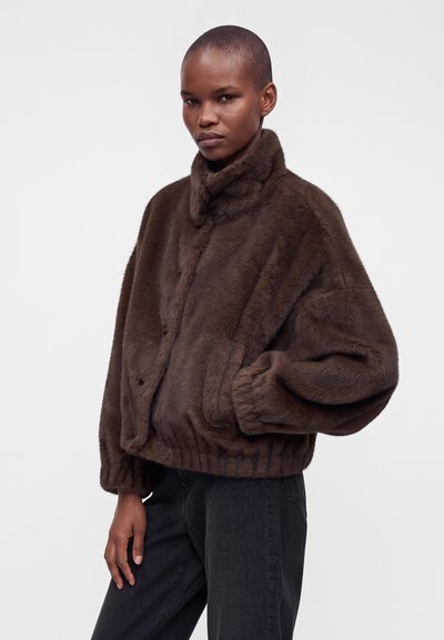 Personne portant une veste en fausse fourrure marron foncé à col montant et un pantalon noir, debout avec une main dans la poche devant un fond uni.