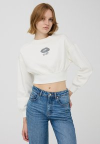 Biała krótka bluza z okrągłym dekoltem i bufiastymi długimi rękawami, z grafiką ust i tekstem. Połączona z niebieskimi spodniami o wysokim stanie.