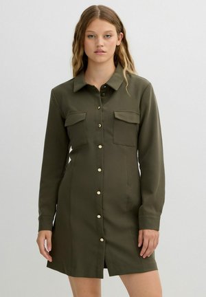 Vestido camisa verde oliva en tejido suave, con un botón delante, dos bolsillos en el pecho y mangas largas con puños de botón.