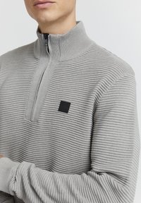 Grå, zip-up genser med ribbestruktur, stående krage, og en liten svart logoetikett på brystet.
