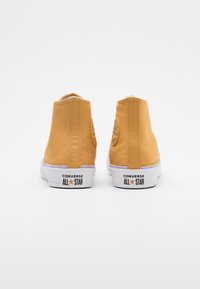 Żółte płócienne trampki typu high-top z białymi gumowymi podeszwami i okrągłą łatą z logo na zewnętrznej kostce. Gładka tekstura, minimalne detale.