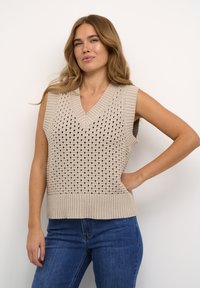 Top sans manches en tricot beige avec un décolleté en V, présentant des motifs en tricot ouvert et un ourlet côtelé. Associé à un jean en denim bleu.