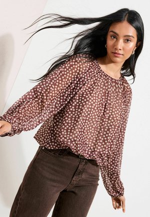 Pruun läbipaistev pluus nõgusa kaelusega ja pikkade puksivarrukatega, millel on heledates toonides täpiline muster. Sobitatud tumepruunide pükstega.