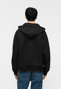 Άτομο εμφανίζεται από πίσω φοράει μαύρο φούτερ με κουκούλα και μπλε τζιν με μεγάλες τσέπες πάνω σε απλό ανοιχτό φόντο.