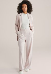 Žena kovrčave kose u blijedo ružičastom odijelu, bijeloj teksturiranoj majici i bijelim tenisicama, stoji s rukama u džepovima na jednobojnoj pozadini.