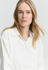 Bijela košulja na dugme s teksturiranim materijalom, kragnom i jednim džepom na prsima. Model ima dugu, ravnu kosu. Meko, prirodno osvjetljenje.