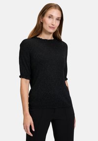 Sort kortærmet sweater med en tekstureret, glitrende finish og en volang i halsudskæringen, parret med sorte bukser. Enkel, tætsiddende design.