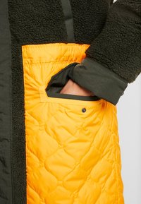 Vadderad jacka med en mörkgrön fleecekropp och en kviltad orange ficka med tryckknapp. Texturerad tyg kontrasterar med glatt material.