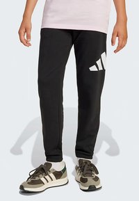 Crne trenirke od mekog materijala s velikim bijelim logotipom na lijevom bedru, uparene s višebojnim sportskim cipelama.