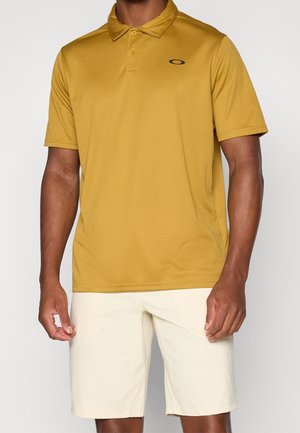 Homme portant un polo à manches courtes jaune moutarde et un short beige clair, debout devant un fond uni clair.