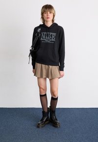 Musta huppari, jossa on "NIKE"-logo, beige laskostettu hame, mustat polven ylle ulottuvat sukat ja mustat nauhakengät keltaisella ompelulla.