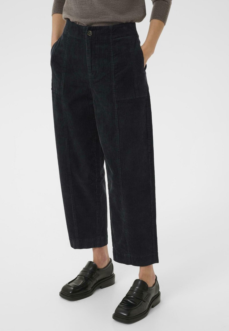 Donker corduroy wijde broek met een subtiele ribbelstructuur, voorzien van zijzakken en een sluiting met een knoop aan de voorkant. Gecombineerd met zwarte loafers.