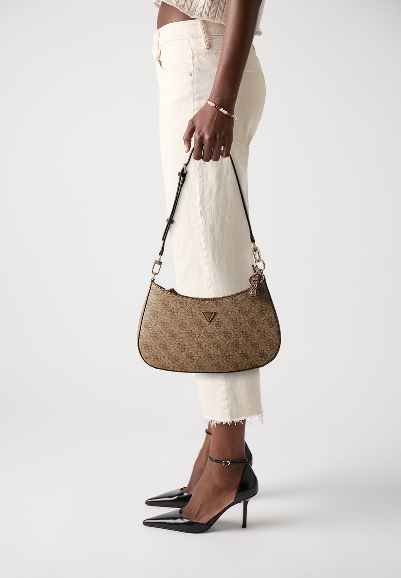 Personne portant un pantalon crème et des talons hauts noirs tenant un sac à épaule marron à motif avec une sangle noire et des accessoires dorés.