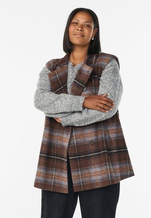 Brown plaid vest with a structured design, featuring gray knit sweater underneath. Notable buttons and layered texture. Dark jeans complete the look.