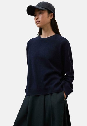 Jersey de punto azul marino con cuello redondo, mangas largas y dobladillo acanalado, combinado con una falda plisada verde oscura y una gorra oscura.