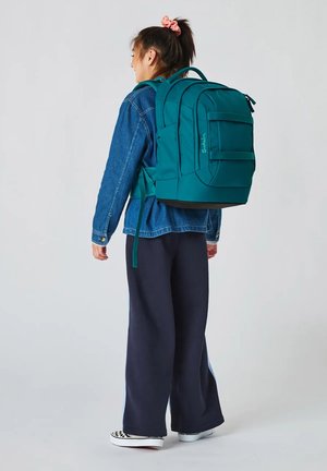 Junge Person mit einem großen türkisen Rucksack, Jeansjacke, weiten dunkelblauen Hosen und karierten Sneakers, die vor einem einfarbigen Hintergrund steht.