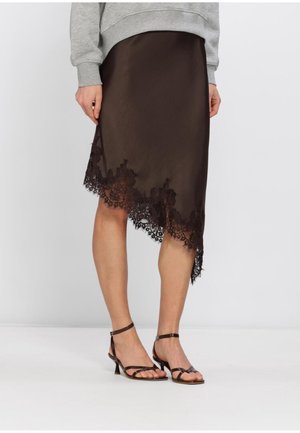 Vrouw draagt een bruine asymmetrische rok met kanten rand, een grijze trui en zwarte sandalen met lage hakken en bandjes, staande op een witte vloer.