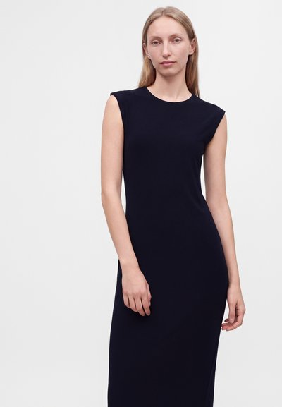 Femme aux longs cheveux blonds portant une robe noire sans manches sur un fond gris clair uni.