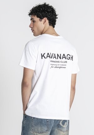 Jonge man met krullend haar, die een wit T-shirt draagt met de tekst "KAVANAGH WINNERS CLUB" op de rug en vervaagde blauwe spijkerbroek.