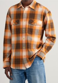Camicia a quadri arancione, marrone e grigia; presenta due tasche sul petto e una vestibilità rilassata con una trama intrecciata.