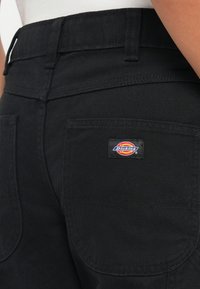 Črne platnene hlače z oprijetim pasom, dvema zadnjima žepoma in vidno oznako blagovne znamke Dickies, prišito na žepu na desni strani.