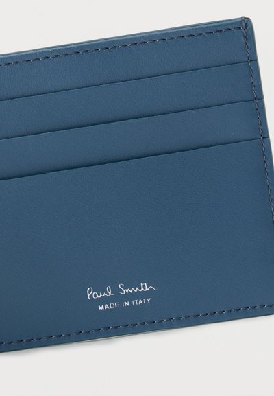 Μπλε δερμάτινη θήκη καρτών με τέσσερις θήκες για κάρτες και λευκή επιγραφή "Paul Smith Made in Italy" στην κάτω δεξιά γωνία.