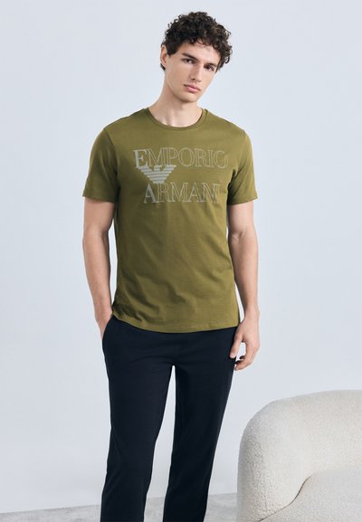 Olivzaļš kokvilnas T-krekls ar "EMPORIO ARMANI" logotipu gaiši pelēkā krāsā, apaļu kakla izgriezumu un īsām piedurknēm, kopā ar tumšām biksēm.
