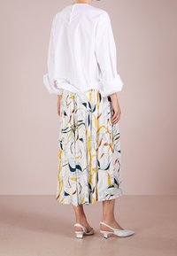 Blusa bianca a cappuccio ampia con apertura sul retro abbinata a una gonna plissettata multicolore a fantasia. Il modello indossa décolleté bianche con cinturino.