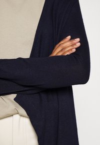 Cardigan lavorato a maglia blu navy con vestibilità ampia, caratterizzato da una texture a coste e un'apertura frontale. Abbinato a un top beige chiaro. Braccia incrociate davanti.