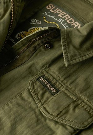 Chaqueta verde oliva con cierre frontal, textura acanalada y un parche con la marca "SUPERDRY". La tela presenta un sutil patrón de espiga.