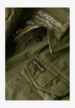 Olivengrøn jakke med lynlås foran, ribbet struktur og et mærke med "SUPERDRY" branding. Stoffet har et diskret hørnmønster.