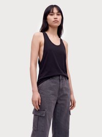Young woman with straight black hair wearing a black sleeveless tank top and gray cargo pants standing against a plain background.