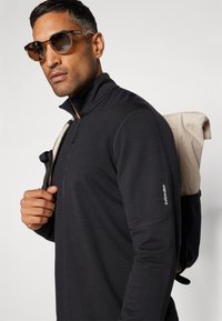 Must be a black zip-up pullover kõrge kaelusega, tekstuurse kangaga ja logoga varrukal. Pruunitoonilised päikeseprillid ja beež seljakott ruudulise mustriga.