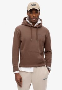 Brązowy bawełniany hoodie z przednią kieszenią i regulowanym kapturem, nałożony na biały t-shirt. Beżowa czapka z logo "Superdry Athletics".