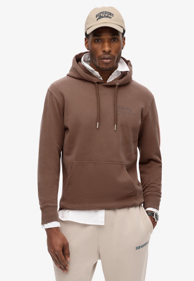 Brązowy bawełniany hoodie z przednią kieszenią i regulowanym kapturem, nałożony na biały t-shirt. Beżowa czapka z logo "Superdry Athletics".