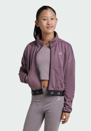 Chaqueta corta morada con cuello alto y bolsillos frontales, con una banda negra en la cintura que presenta patrones triangulares, combinada con leggings y una camiseta grises.