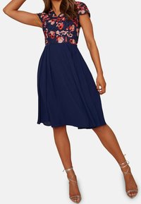 Tummansininen, polvipituinen mekko, jossa on lyhyet hihat, kirjailtu kukkakuviointi yläosassa ja liehuva hame. Yhdistetty hopeisiin remmiavokkaisiin.