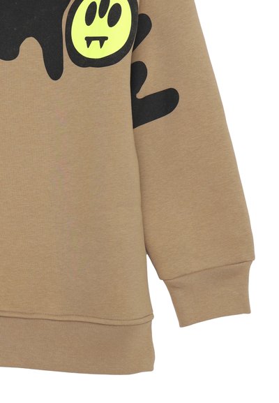 Manica di felpa color tan con un design di vernice nera che scorre e una faccina sorridente gialla con canini da vampiro sulla parte superiore del braccio.