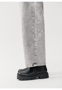 Botas negras hasta el tobillo con suela gruesa, parte superior de cuero suave y punta redonda, combinadas con jeans grises que presentan un lavado desvanecido y un dobladillo deshilachado.