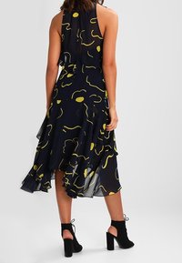 Laivastonsininen mekko, jossa on abstrakteja keltaisia kuvioita, kerroksellisia röyhelöitä ja miehustakotelo. Yhdistetty mustien nilkkahihnoilla varustettujen palikka-korkokenkien kanssa.