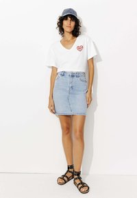 T-shirt blanc à col en V avec un graphique de cœur rouge, jupe en denim bleu clair et sandales noires à lanières avec des clous, se tenant devant un fond blanc.