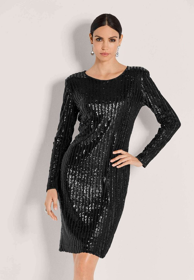 Frau trägt ein enges, schwarzes, mit Pailletten besetztes Kleid mit langen Ärmeln, Hand an der Hüfte, dunkles Haar zurückgebunden, trägt hängende Ohrringe und einen Ring.