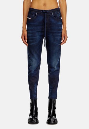 Femme portant un jean jogger bleu foncé avec un cordon et des bottines noires brillantes à bout carré, debout devant un fond uni.