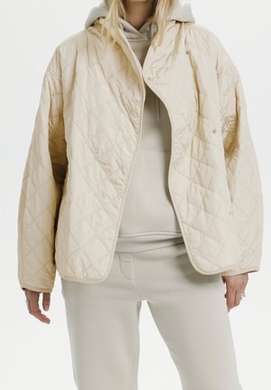 Kobieta ubrana w beżową pikowaną kurtkę na beżowej bluzie z kapturem i pasujących spodniach, stojąca na tle białego tła.