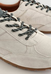 Sneakers in suede grigio chiaro con dettagli in pelle grigio scuro, dotate di lacci neri e bianchi con motivo e una suola in gomma testurizzata.