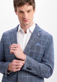 Man past een manchet van een blauw geblokt colbert over een wit overhemd, staande tegen een effen witte achtergrond.