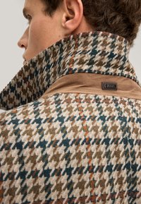 Hahnentrittmuster-Stoff in erdigen Tönen von Grün, Orange und Beige, mit einem braunen Wildlederkragen und einem sicheren Etikett-Detail.