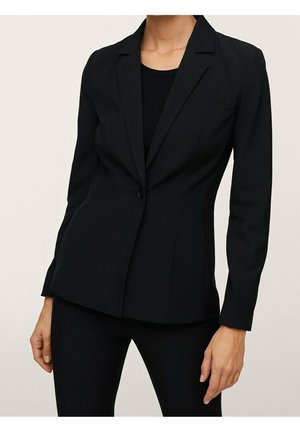 Kvinde iført en tætsiddende sort blazer med en enkelt knap, kombineret med sorte bukser mod en neutral baggrund.