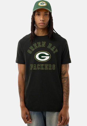 Młoda osoba z długimi włosami, ubrana w czarną koszulkę Green Bay Packers i zieloną czapkę z logo drużyny.